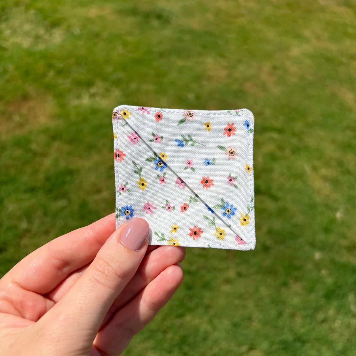 A hand holding a square white fabric patch with a colorful floral print, set against a blurred green grass background.
