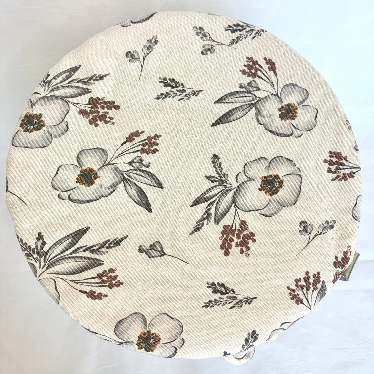 A round fabric-covered stool or cushion with a cream-colored linen-like surface featuring a floral print in gray and brown, photographed from above on a white background.