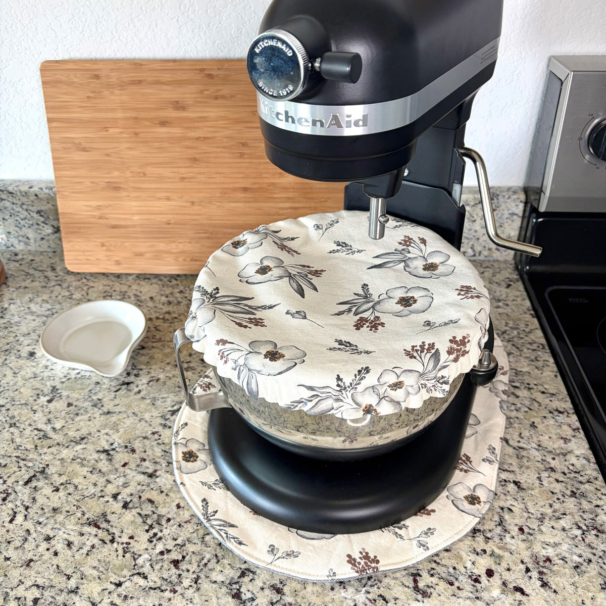 A black KitchenAid stand mixer with a floral-patterned fabric cover on its bowl, set on a granite kitchen countertop next to a wooden cutting board and a small white dish.
