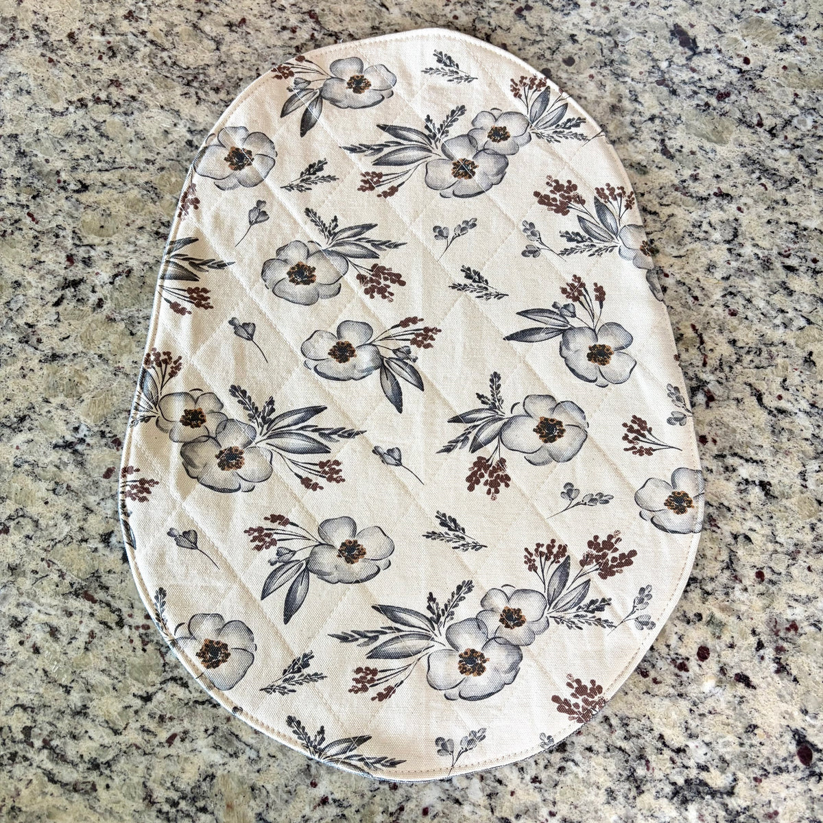 An oval quilted mat with a cream background and a floral print featuring gray and brown flowers, leaves, and berries, laid flat on a speckled granite countertop. Bowl Lift Base