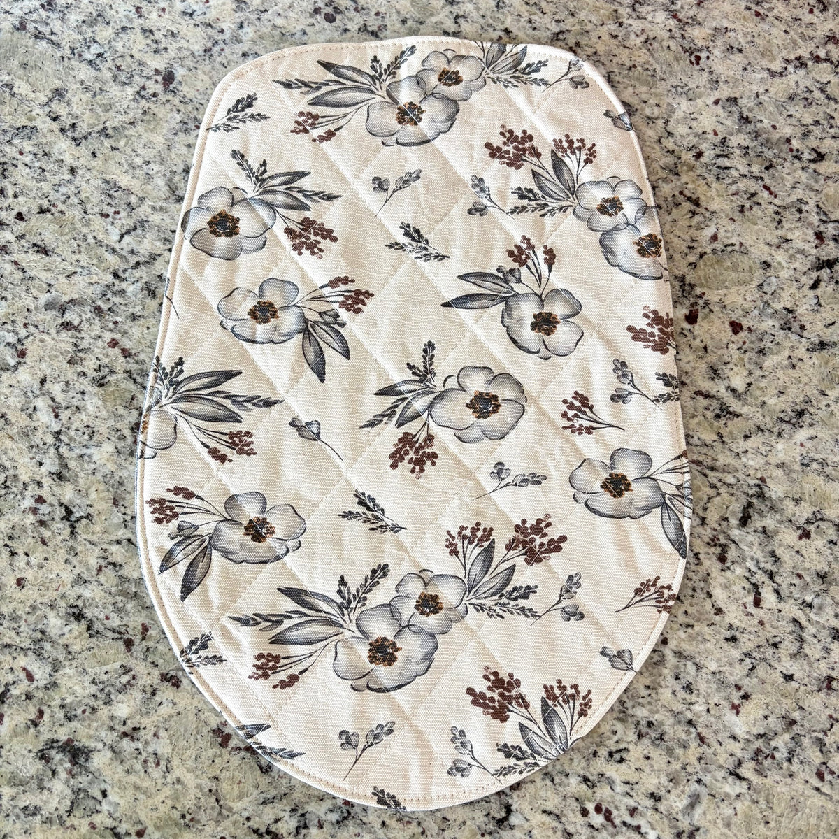 An oval quilted fabric mat in the shape of a head tilt stand mixer's base, featuring a floral print on a cream background. The print includes gray and brown flowers, leaves, and berries.