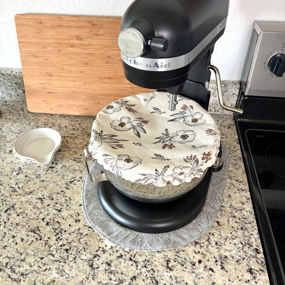 A black KitchenAid stand mixer with a floral-patterned fabric cover on its bowl, set on a gray mat on a granite kitchen countertop next to a wooden cutting board and stove.