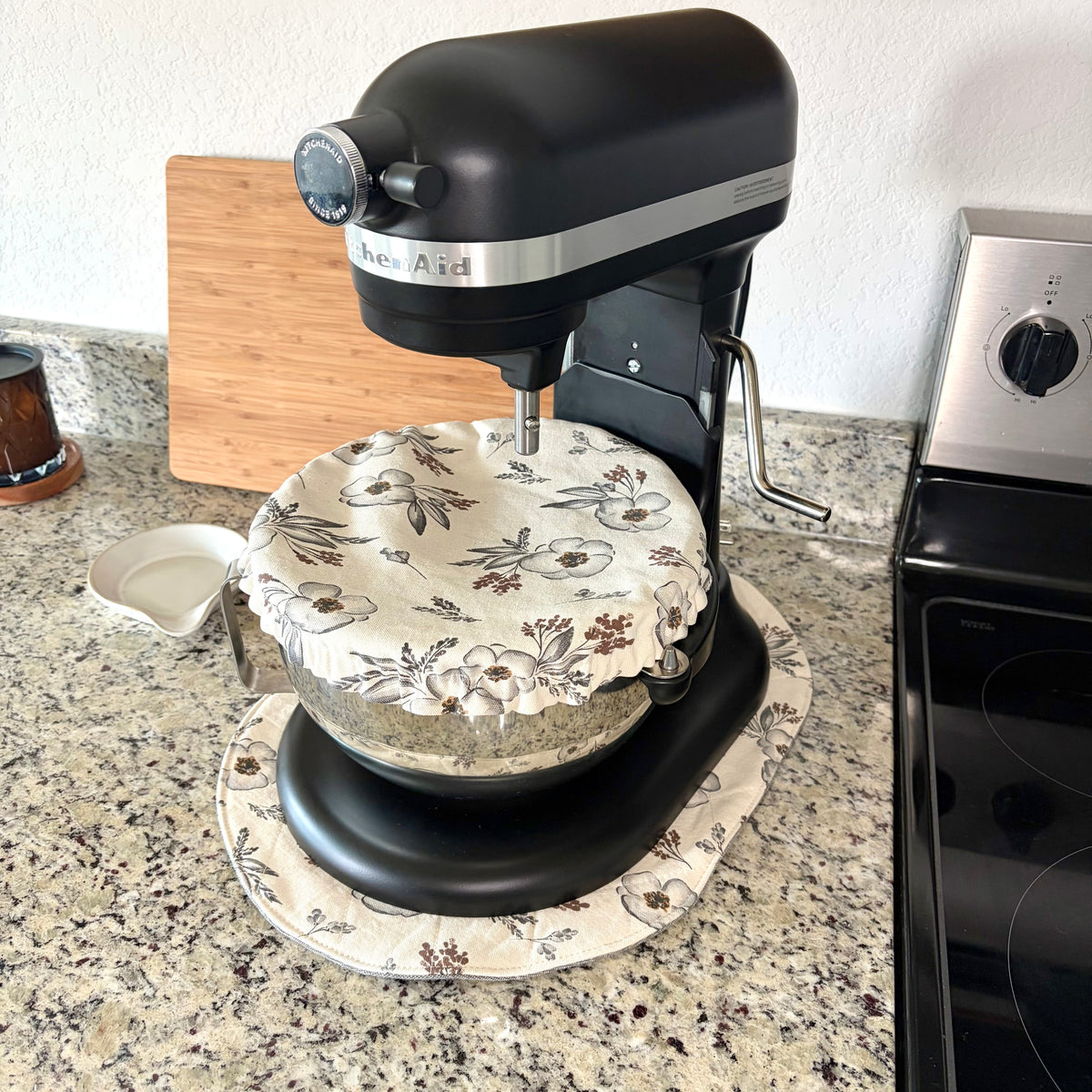 A KitchenAid stand mixer with a black body and silver band, set on a granite kitchen counter next to a stove. The mixer bowl is covered with a cream-colored fabric dust cover featuring a brown and beige floral pattern. ITs sitting on a matching fabric mat.