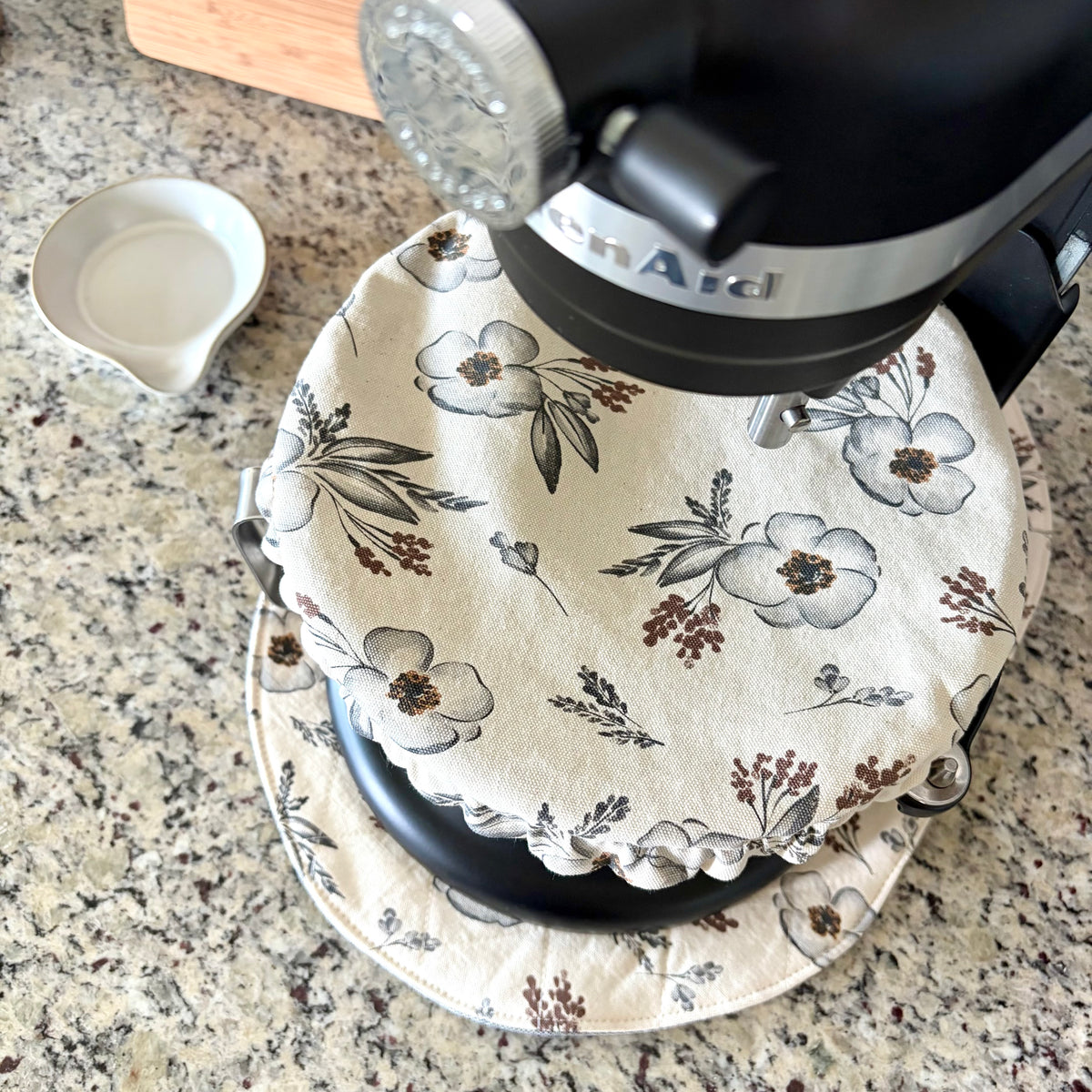 A floral kitchen bowl cover with a ruffled edge draped over the base of a black stand mixer on a granite countertop.