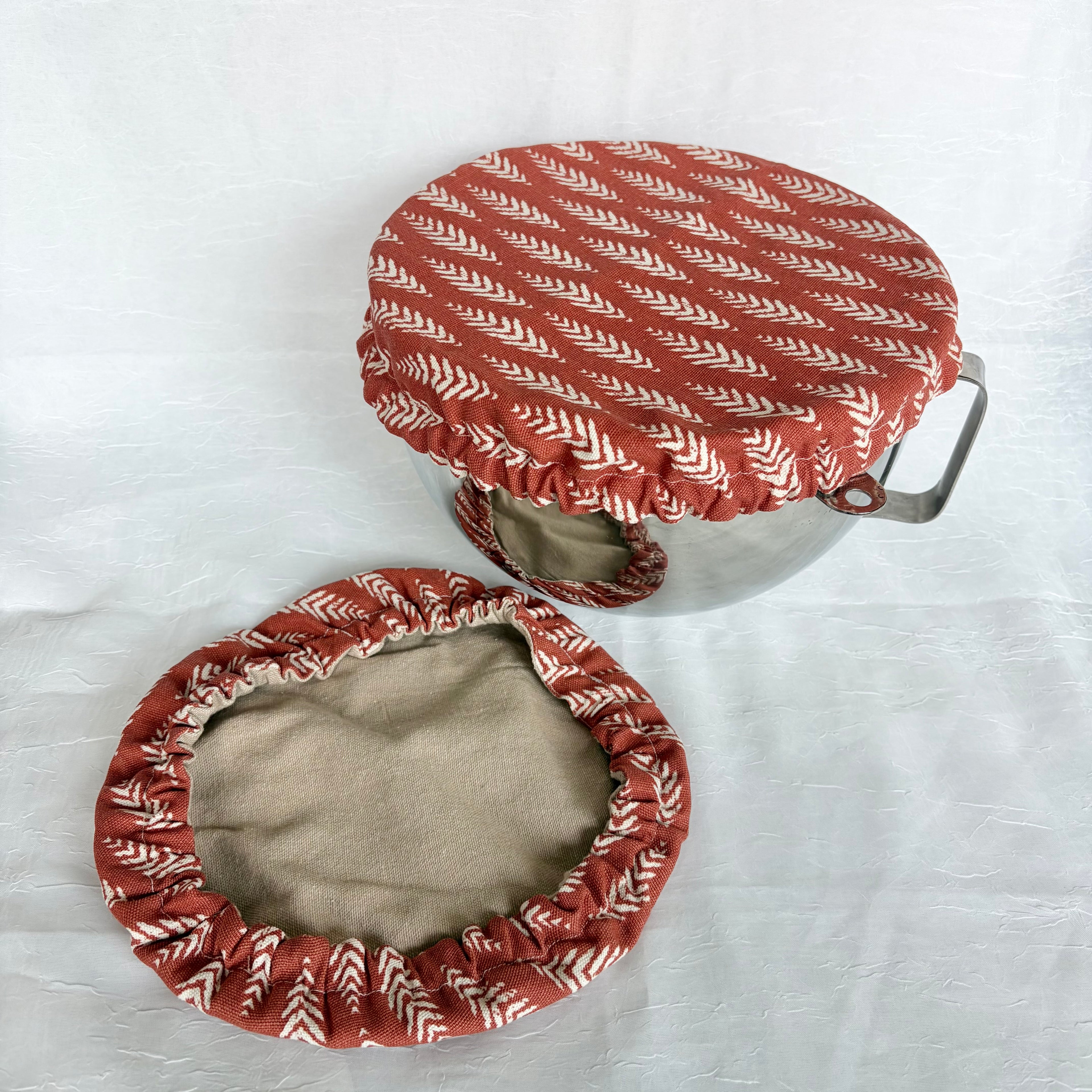 Dark Orange with white floral design in a vertical patterned bowl cover on a metal bowl with a handle on a white background with another bowl cover upside down on the table showing the tan side