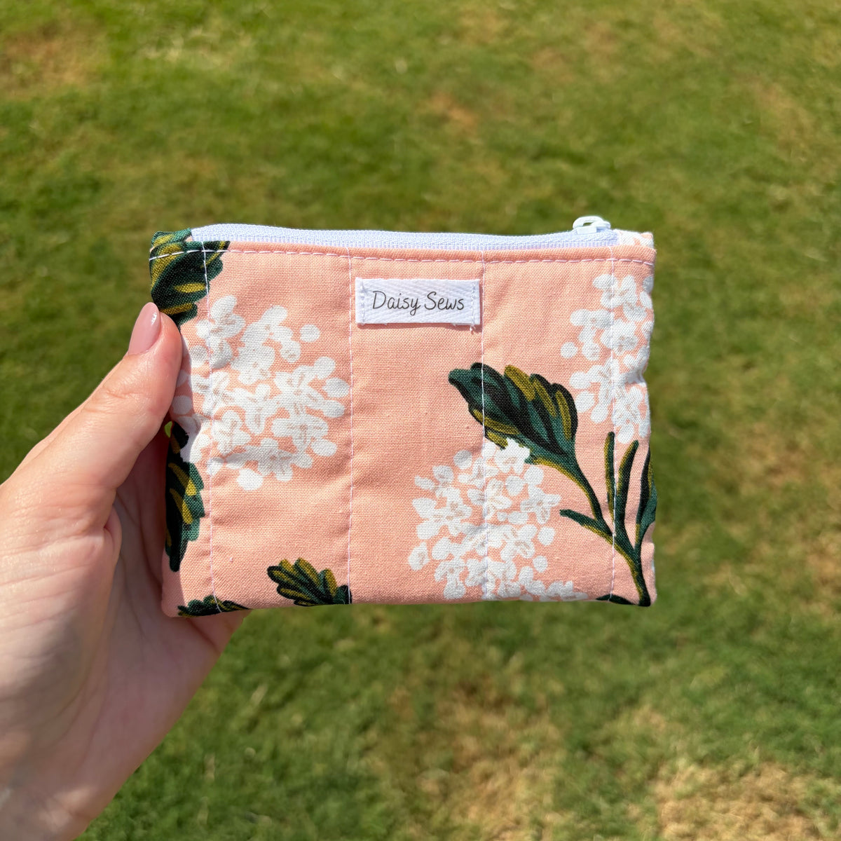 A hand holding a small zippered pouch with a pink floral print, white flowers, and green leaves, set against a blurred green grass background. The pouch has a white label that reads 'Daisy Sews'.