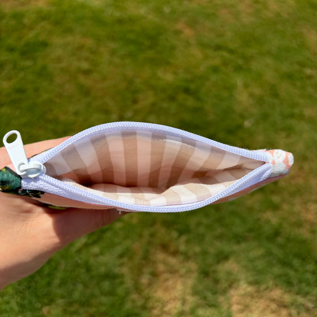 A hand holding open a small zippered pouch outdoors on a grassy lawn. The pouch has a white zipper, a beige and white striped interior, and a small green floral fabric tab attached to the zipper pull.