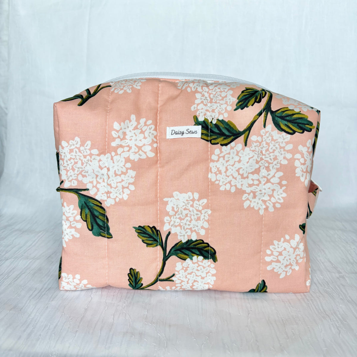 A quilted cosmetic bag in a peachy pink fabric with a white floral and green leaf print, featuring a small white label that reads 'Daisy Sews'.