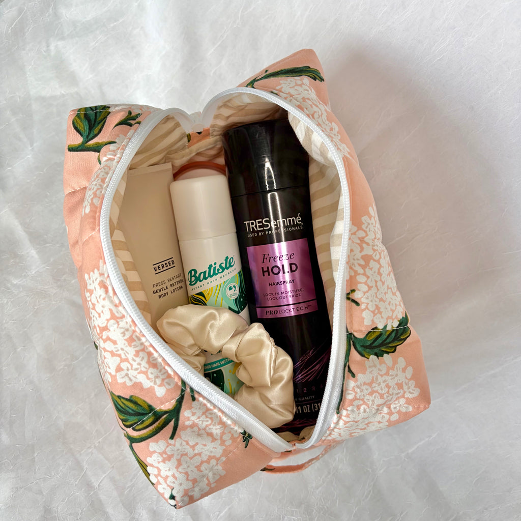 An opened toiletry bag with a pink floral print and white zipper, filled with personal care items including a TRESemmé hairspray, Batiste dry shampoo, and a cream scrunchie, laid on a white textured fabric background.