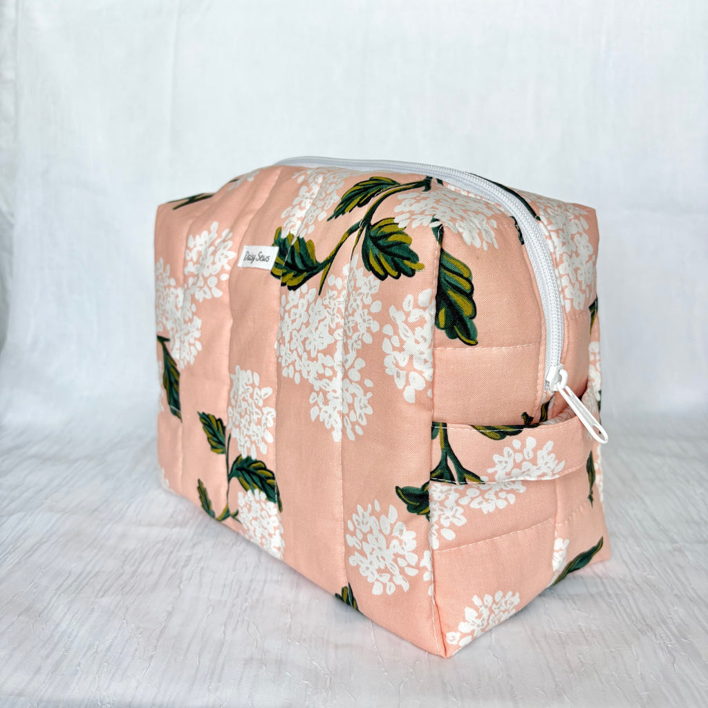 A quilted cosmetic pouch with a peach-pink floral print featuring white flowers and green leaves, shown on a light gray fabric background.