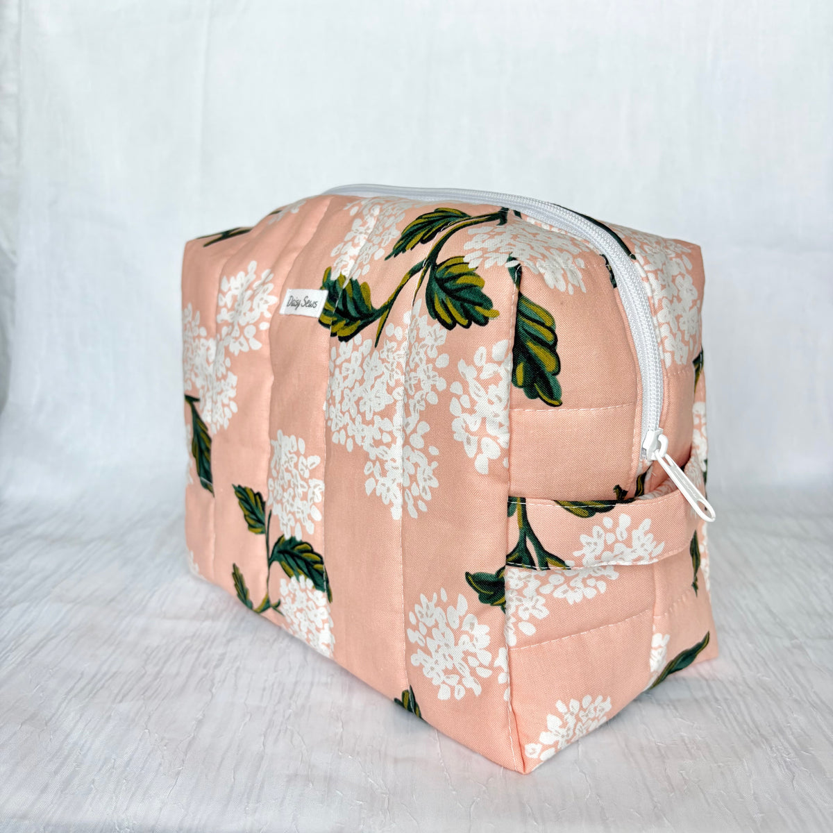 A quilted cosmetic pouch with a peach-pink floral print featuring white flowers and green leaves, shown on a light gray fabric background.