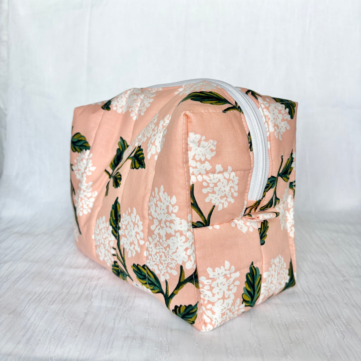A rectangular zippered pouch with a peach-pink fabric cover featuring a white floral print and green leaves, shown on a white textured fabric background.