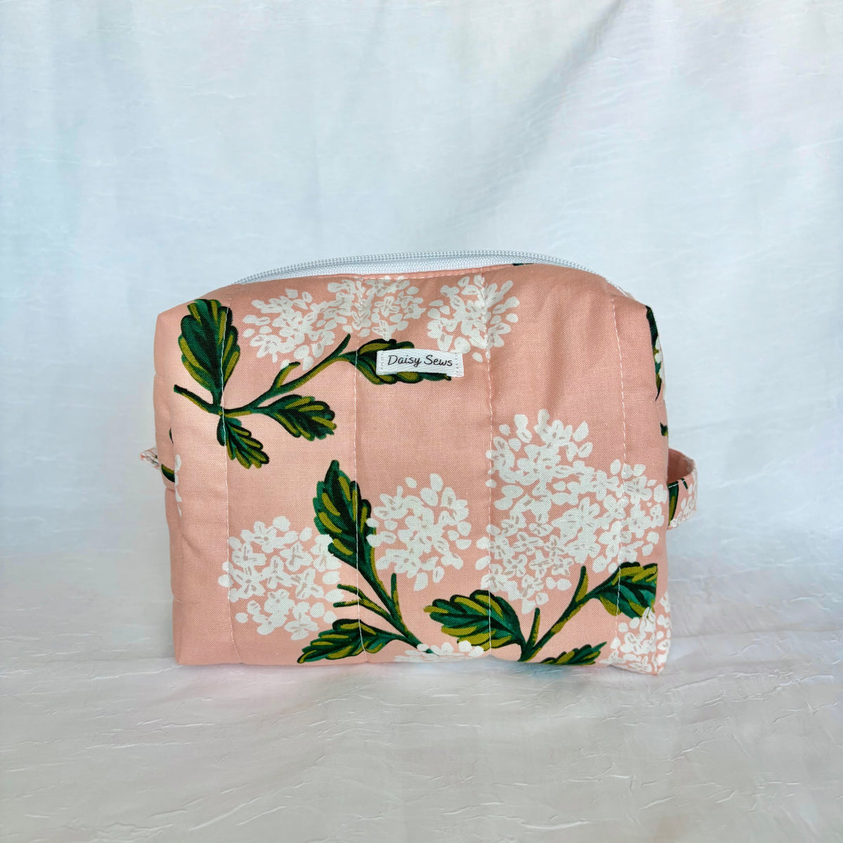 A rectangular zippered pouch in a peachy pink fabric with a white floral and green leaf pattern, featuring a small white label that reads 'Daisy Sews'.