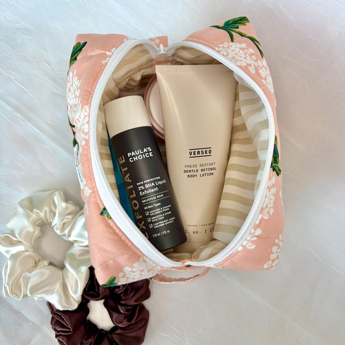 A peach and white floral zippered cosmetic pouch filled with skincare products, including a black Paula's Choice body wash bottle and a beige Versed body lotion tube, arranged on a white textured fabric background with satin scrunchies.