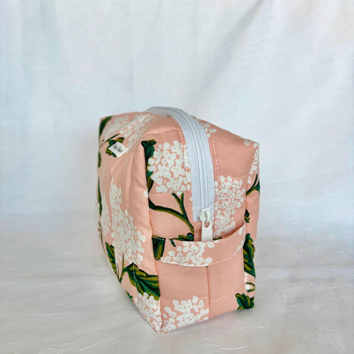 A square zippered pouch with a peach-pink background and a white floral print, featuring a white zipper and a small fabric tag, set against a white fabric backdrop.