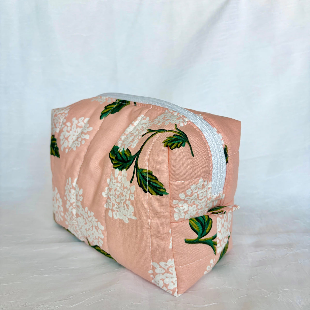 A quilted cosmetic pouch in a peachy pink fabric with a white floral pattern and green leaves, featuring a silver zipper closure, set against a white fabric backdrop.