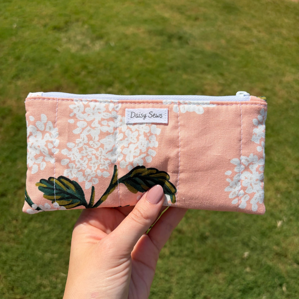 A hand holding a small quilted zip pouch with a pink and white floral pattern and green leaf accents, set against a blurred green grass background. The pouch features a white label reading 'Daisy Sews'.
