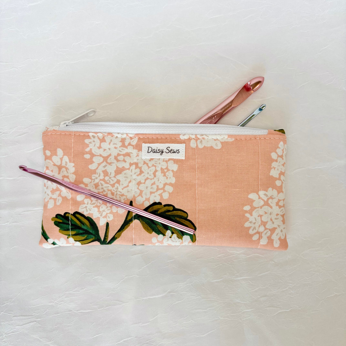 A fabric zippered pouch with a peach background, white floral print, and green leaves, featuring a small 'Daisy Sews' label. Two crochet hooks are tucked into the top zipper.
