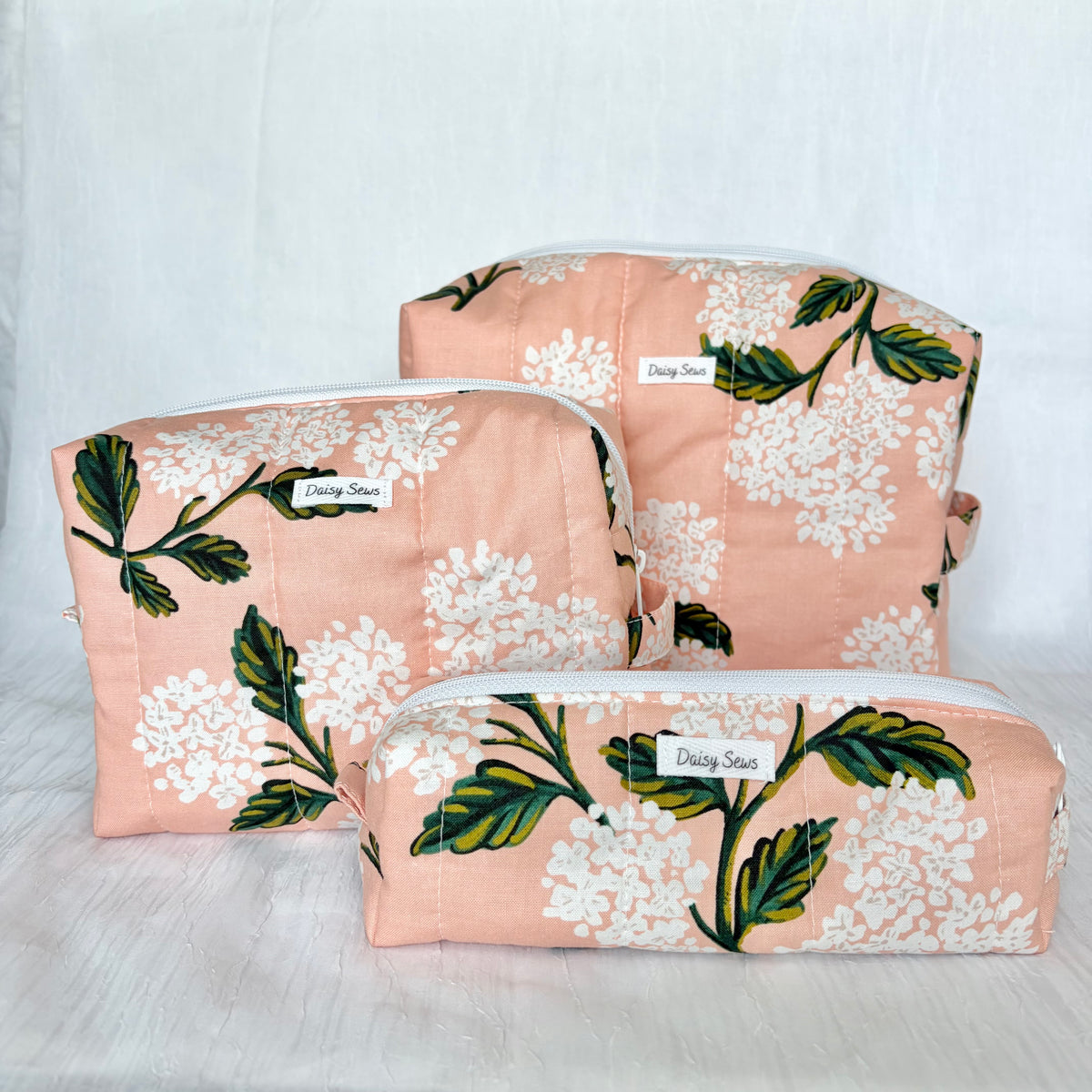 Three fabric cosmetic pouches in a matching set, featuring a peach background with a white floral and green leaf pattern, arranged on a light gray fabric backdrop.