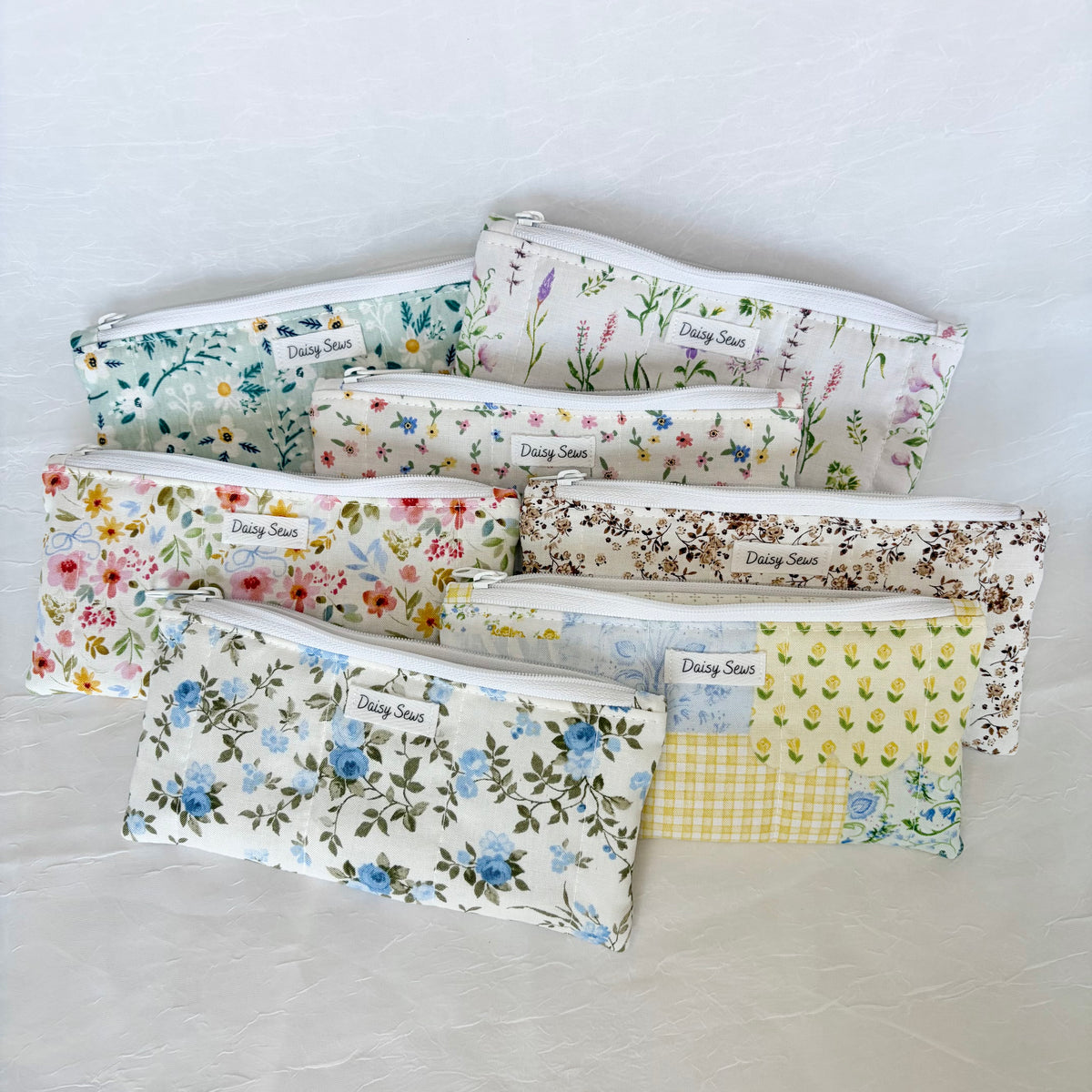 A set of seven fabric zippered pouches with white zippers, arranged in a fan shape on a light gray textured background. Each pouch features a different floral, botanical, or gingham pattern, and they all have a small white fabric label sewn onto the front that reads 'Daisy Sews'.
