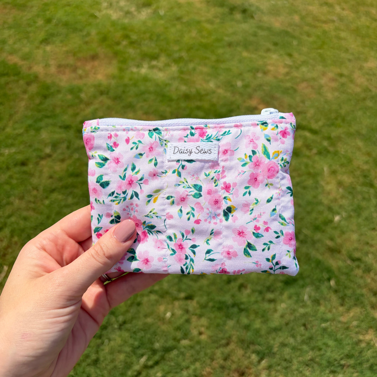 A hand holds a small quilted pouch with a pink and green floral print against a blurred green grass background. The pouch has a white label that reads 'Daisy Sews'.