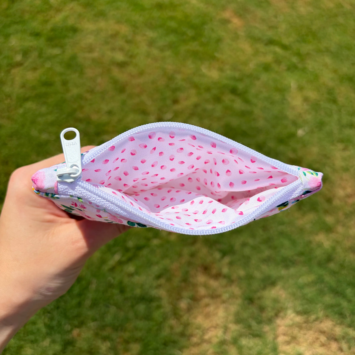 A hand holding an open zippered pouch with a pink floral print exterior and a light pink interior dotted with small pink petals, set against a blurred green grass background.