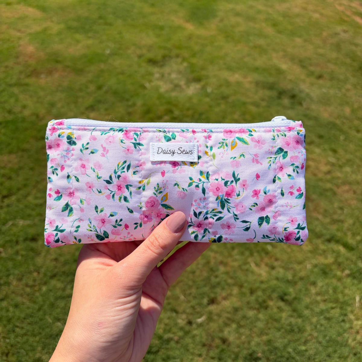 A hand holding a pink floral zippered pouch outdoors against a blurred green grass background. The pouch has a white label that reads Daisy Sews.