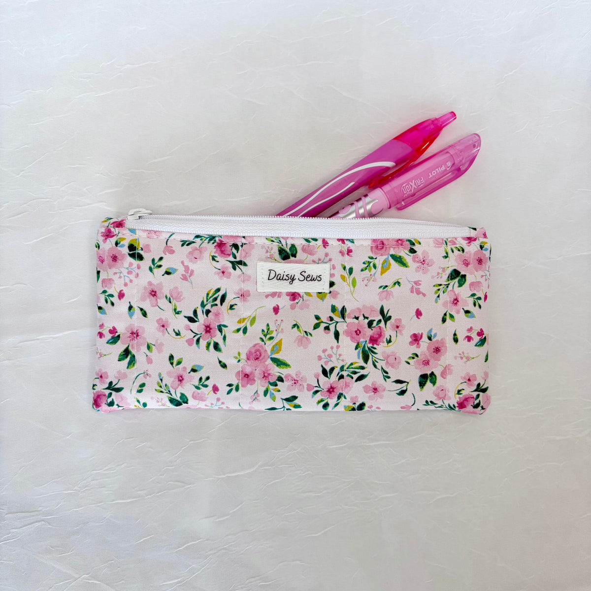 A fabric pencil case with a pink and green floral pattern, featuring a white label that reads 'Daisy Sews'. Two pink pens are tucked inside the zippered case, which is laid flat on a textured white surface.