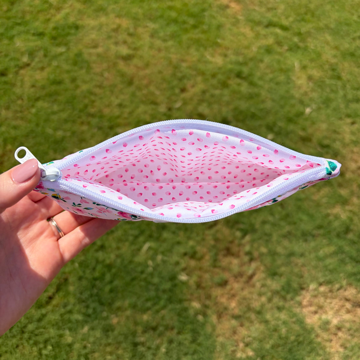 A hand holds an open zippered pouch with a pink and white petal print, set against a blurred green grass background.
