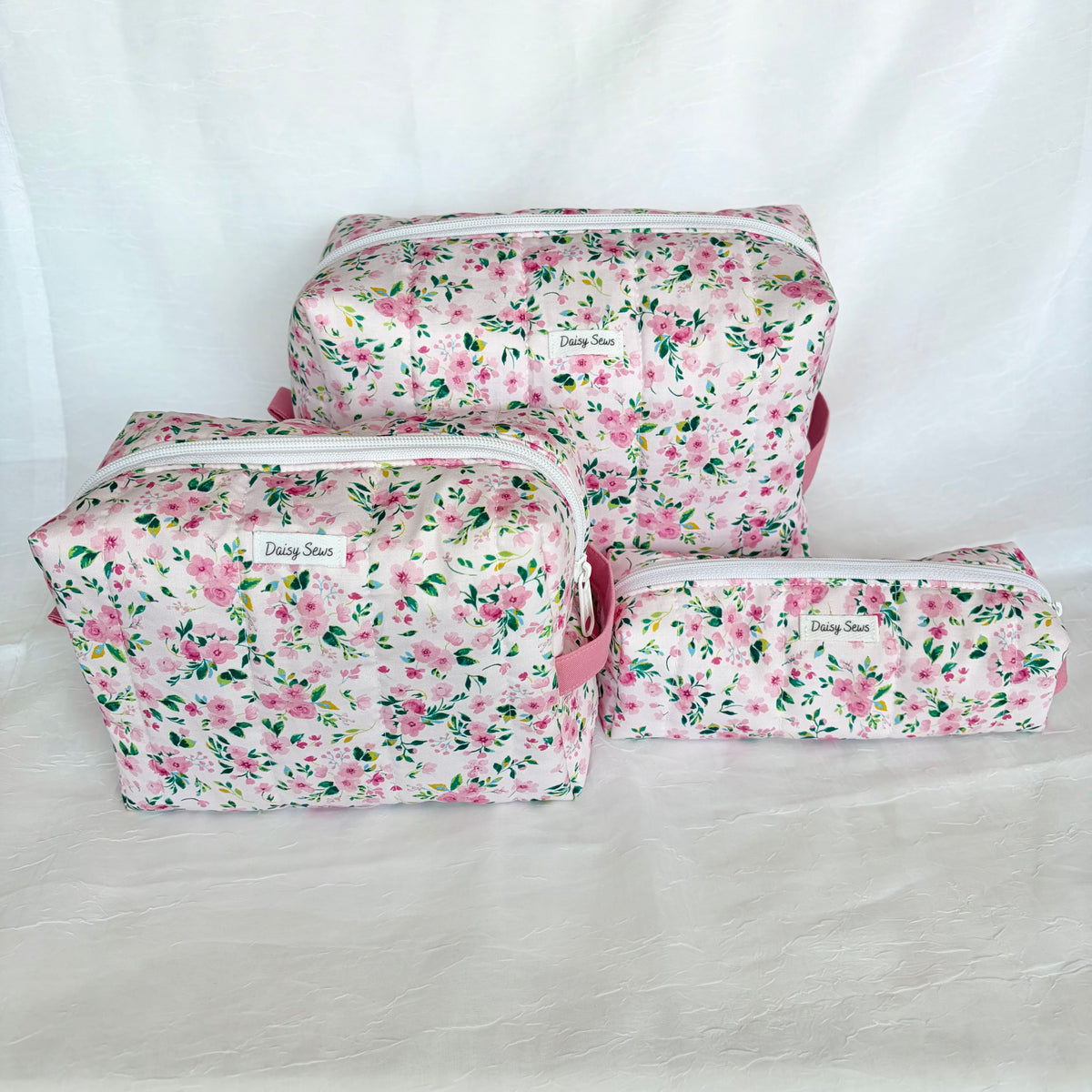 Three floral makeup bags in different sizes arranged on a white fabric backdrop. The bags feature a pink and green floral pattern with white zippers and pink handles.