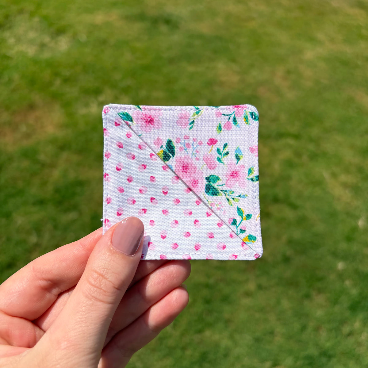 A hand holding a small square fabric patch with a white background, pink floral print, and green leaves, set against a blurred green grass background.