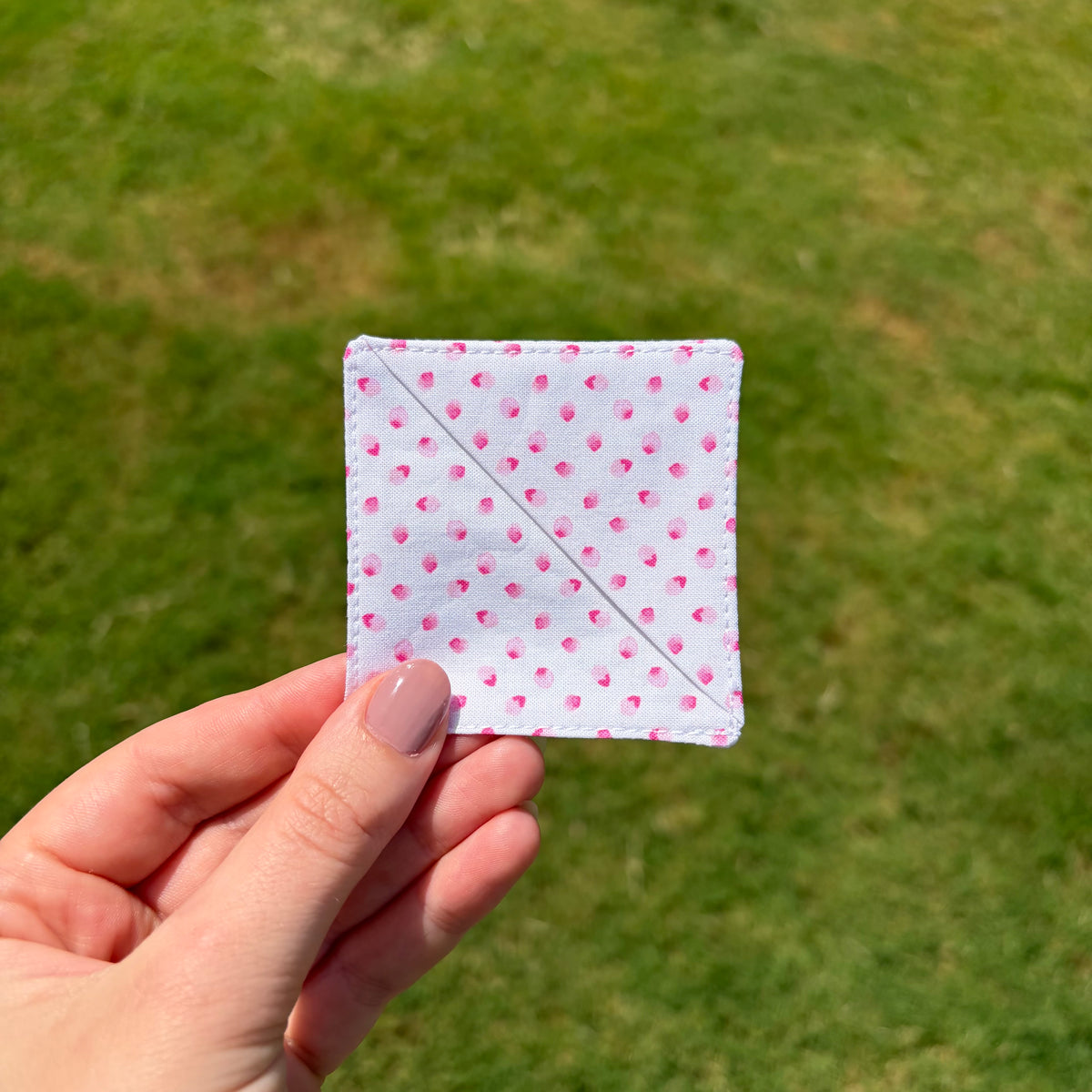 A hand with mauve-painted nails holds a small square white fabric patch with pink petal prints and a diagonal stitched line, set against a blurred green grass background.