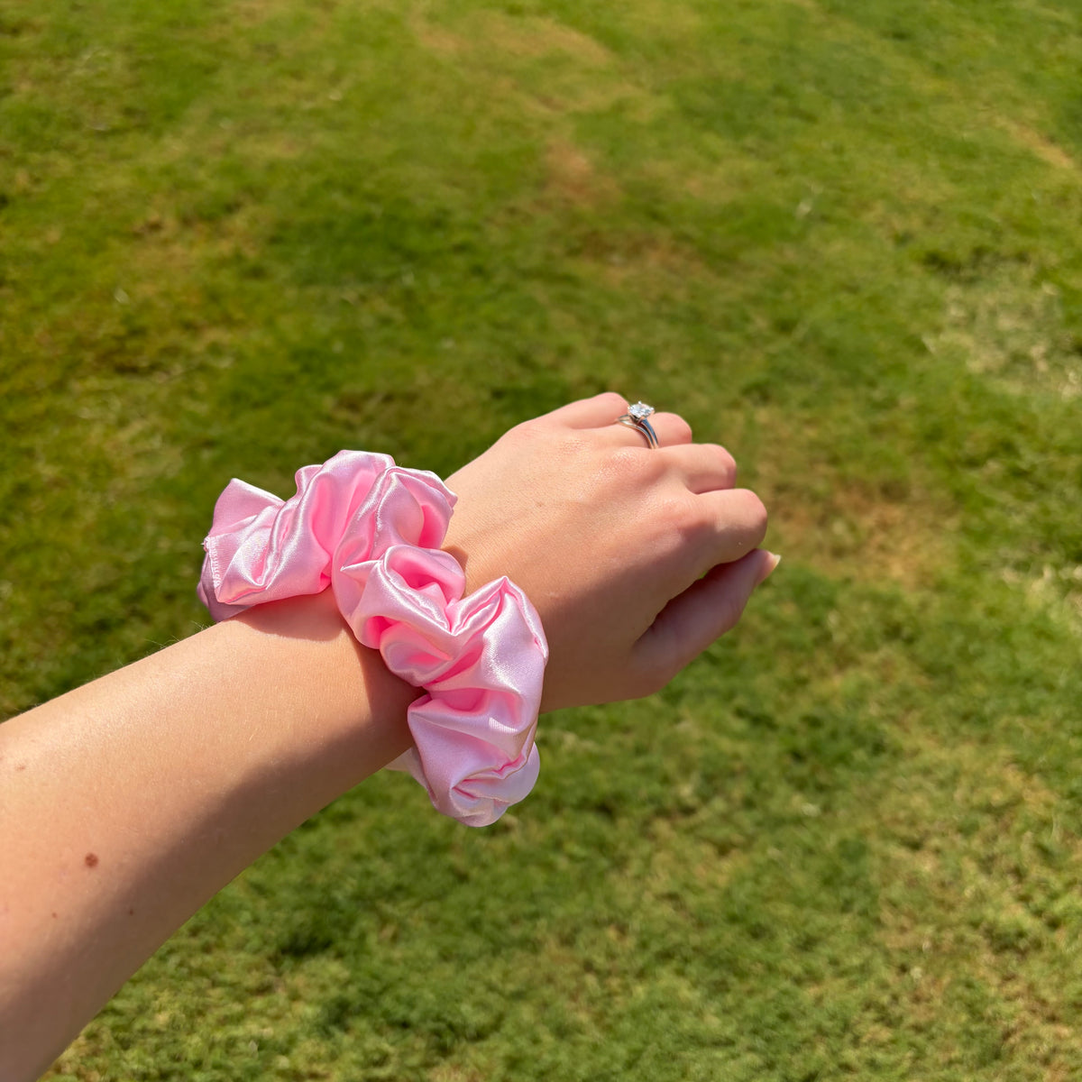A person's wrist wearing two light pink satin scrunchies, photographed outdoors against a blurred green grass background.