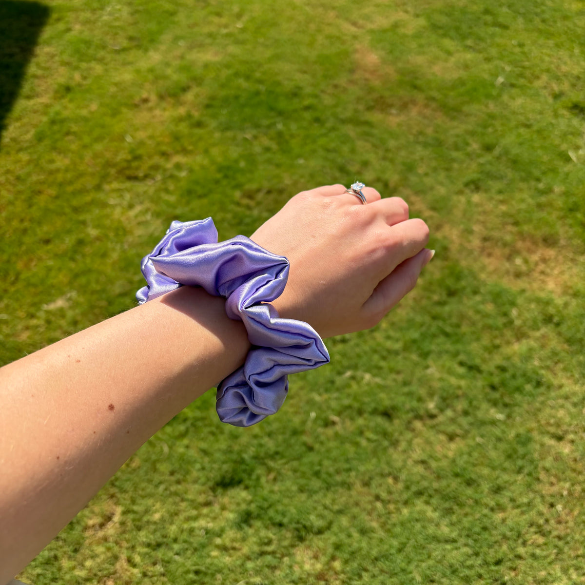 A lavender purple satin scrunchie worn on a person's wrist, with a blurred green grass background.