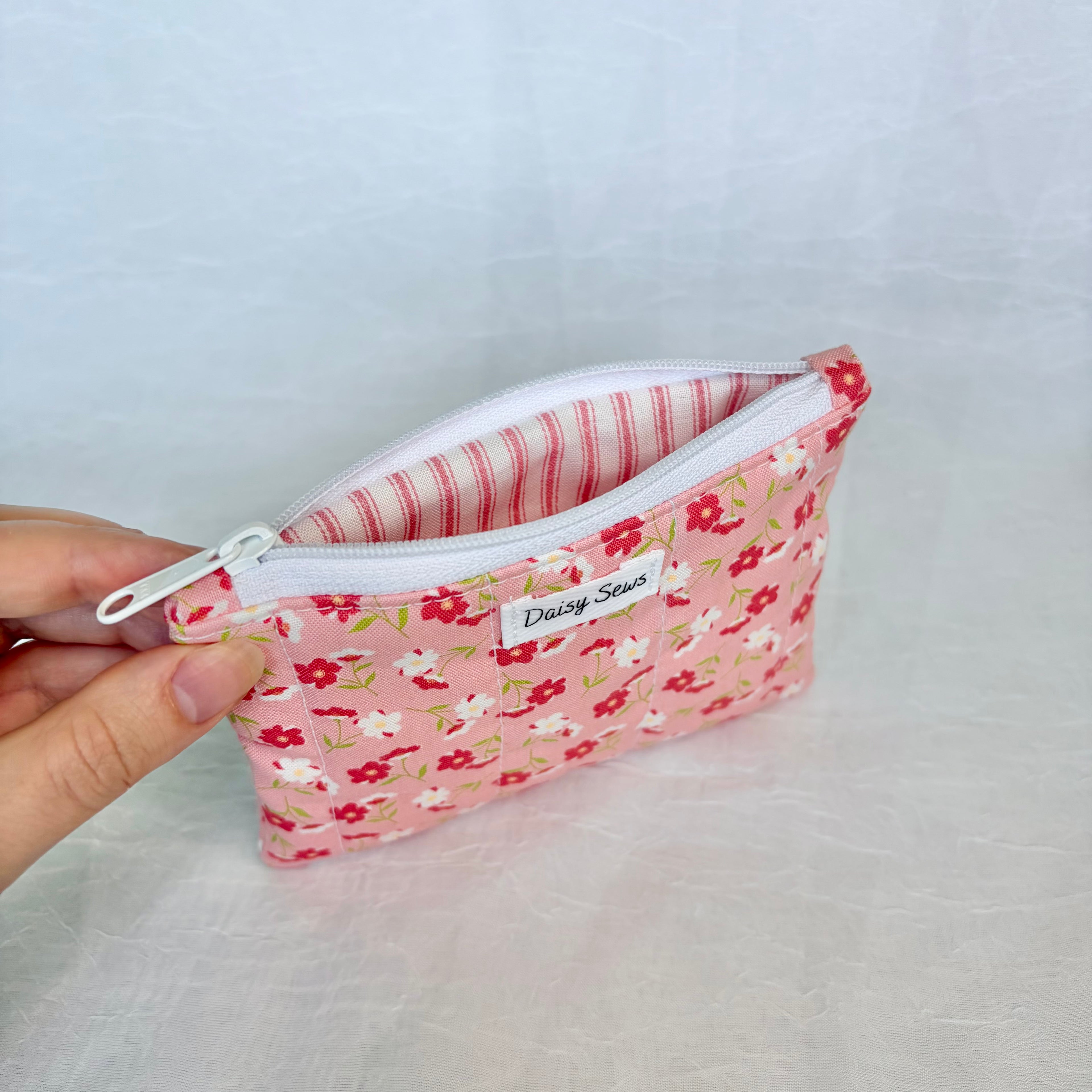 Floral-patterned zipper pouch with pink striped liner held by a hand on a light gray background