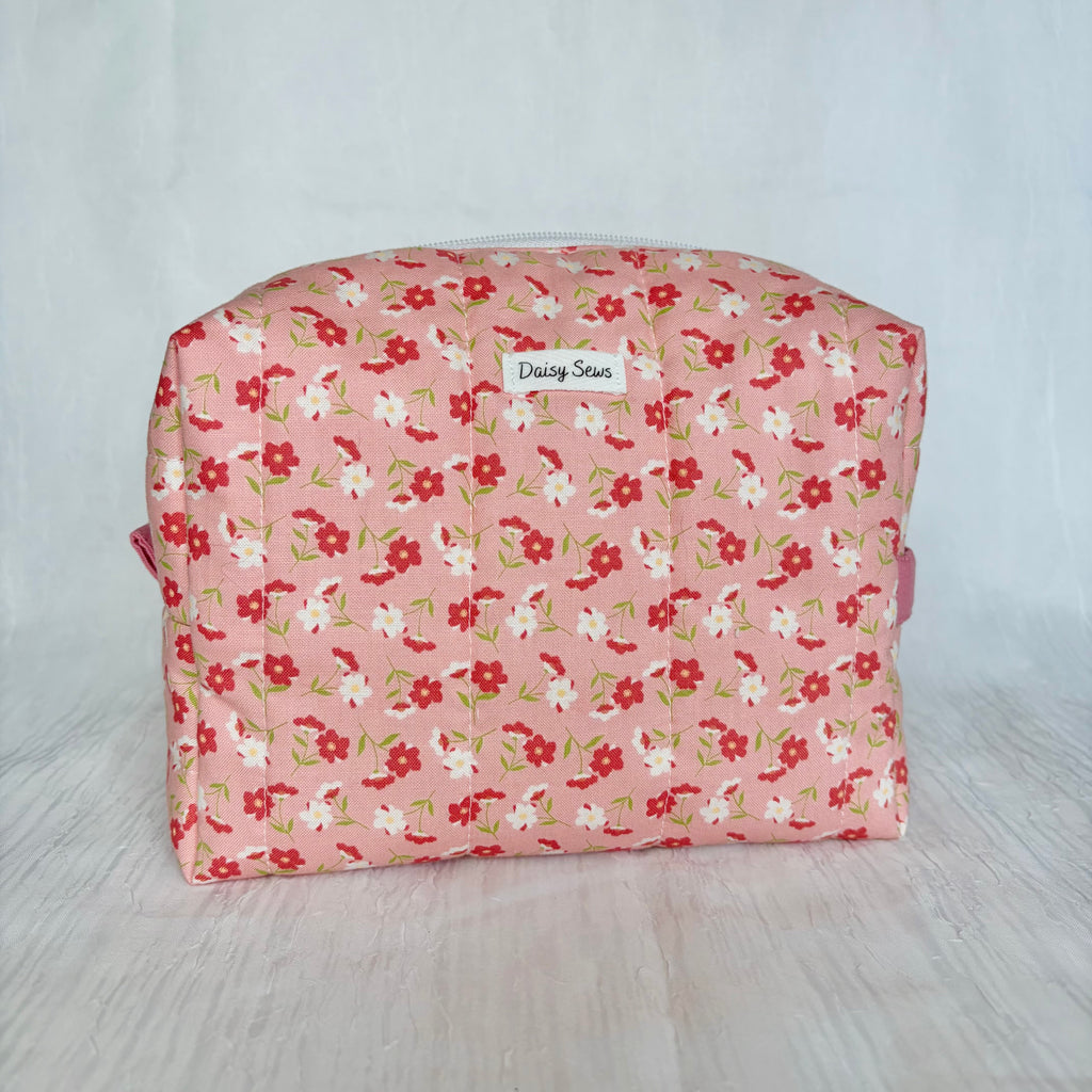 A rectangular cosmetic pouch with a pink floral print, featuring red and white flowers with green stems, and a small white label reading 'Daisy Sews'.