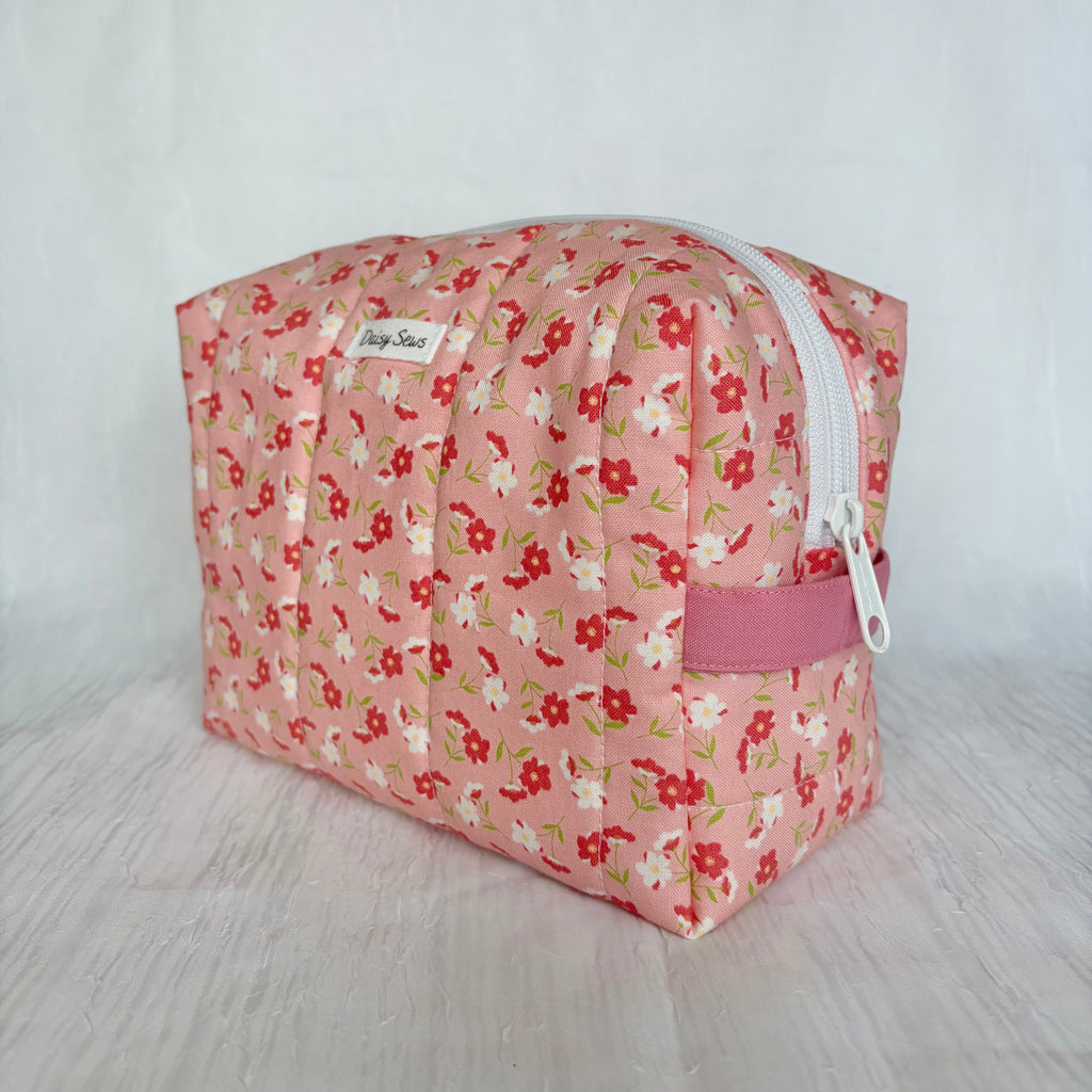 A pink zippered cosmetic pouch with a cherry and white floral pattern, featuring a small white brand label that reads 'Daisy Sews' and a pink zipper pull tab.