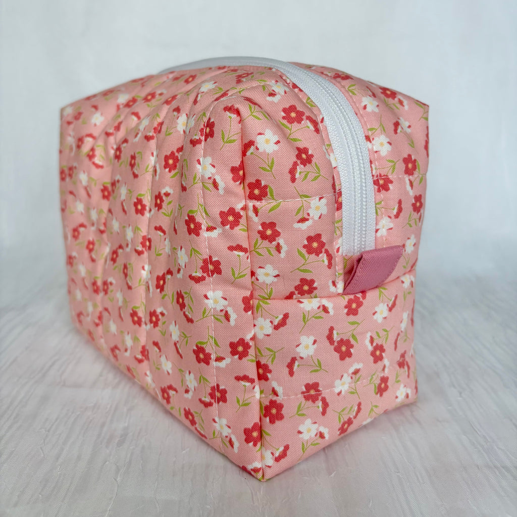 A quilted fabric cosmetic pouch with a pink floral print, featuring red and white flowers with green leaves, a white zipper, and a small pink pull tab, set against a light gray background.