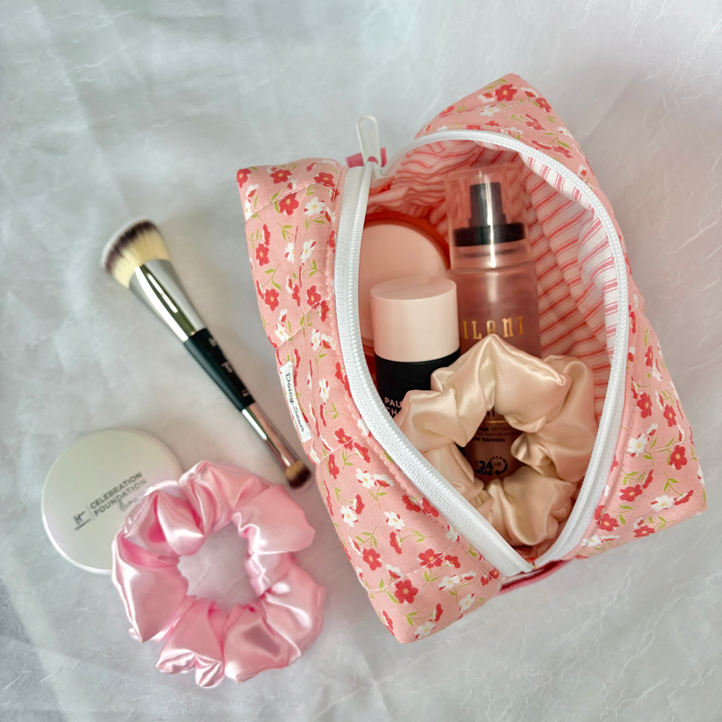 A pink floral makeup bag with a white zipper, shown open with various cosmetics inside, alongside a pink satin scrunchie and a makeup brush on a white textured fabric background.