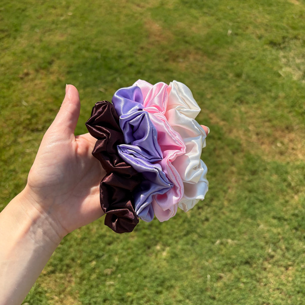 A hand holding a cluster of four satin scrunchies in dark brown, lavender, pink, and white, set against a blurred green grass background.