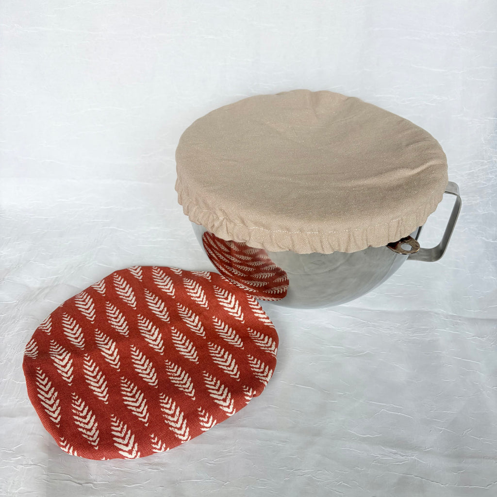 Tan bowl cover on a metal bowl with a handle on a white background with another Dark Orange with white floral design in a vertical patterned bowl cover on the table 