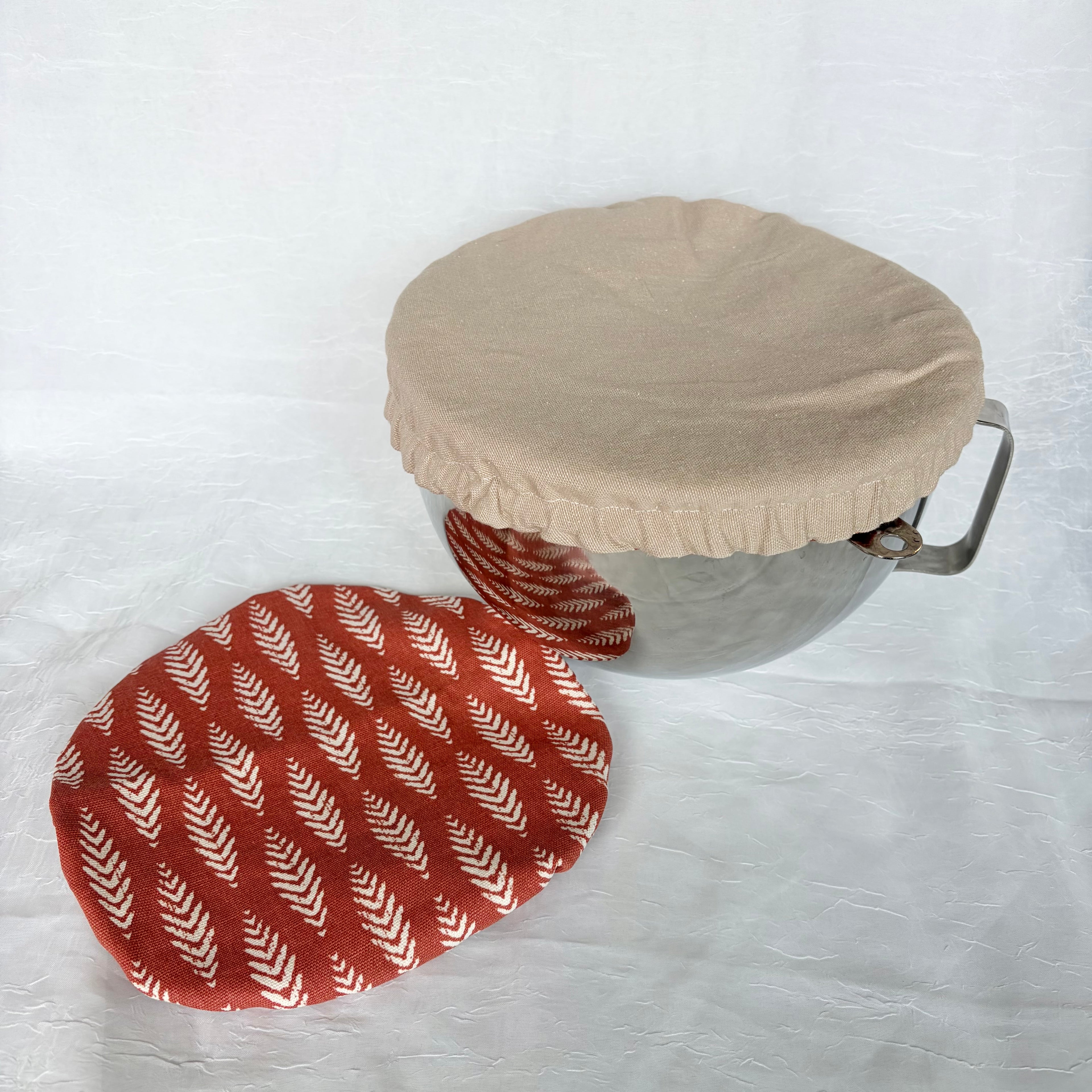 Tan bowl cover on a metal bowl with a handle on a white background with another Dark Orange with white floral design in a vertical patterned bowl cover on the table 