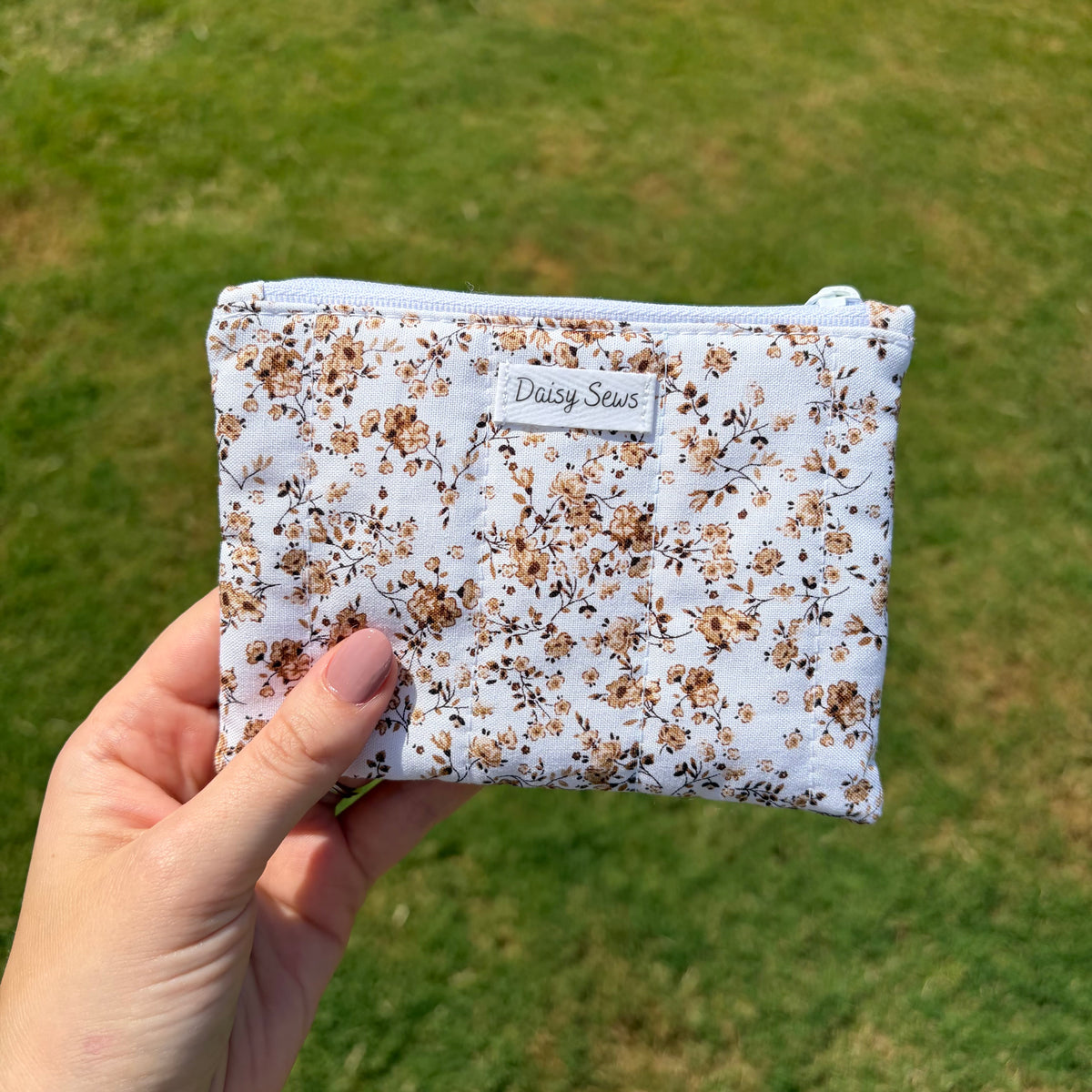 A hand holding a small zippered pouch with a white background and a brown floral print. The pouch has a small white label that reads 'Daisy Sews'.
