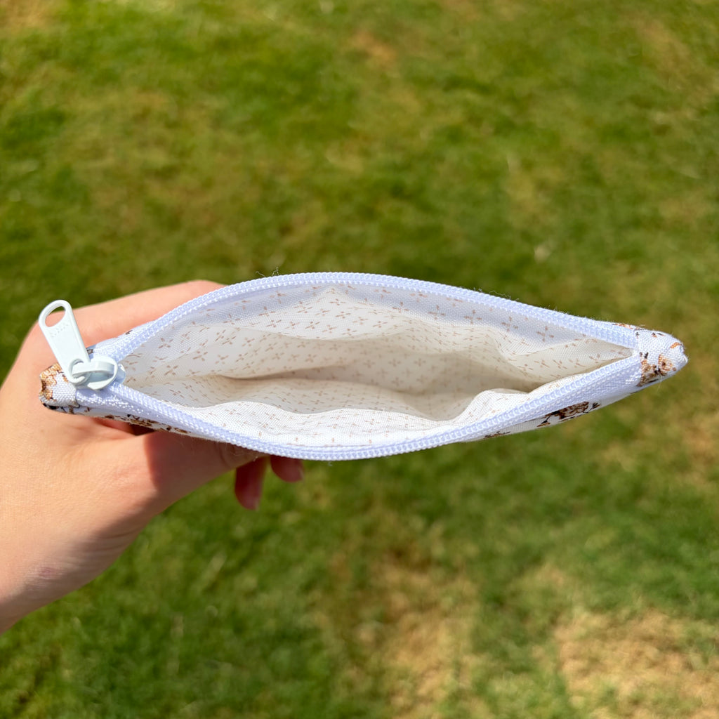 A hand holding an open zippered pouch with a white and brown floral patterned fabric exterior and a light beige dotted lining, set against a blurred green grass background.