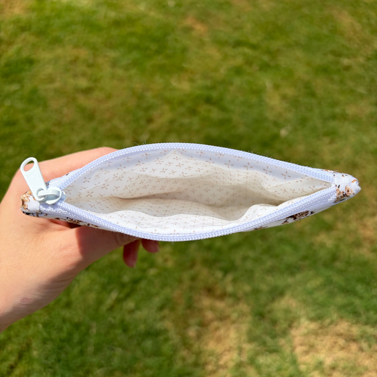 A hand holding an open zippered pouch with a white and brown floral patterned fabric exterior and a light beige dotted lining, set against a blurred green grass background.