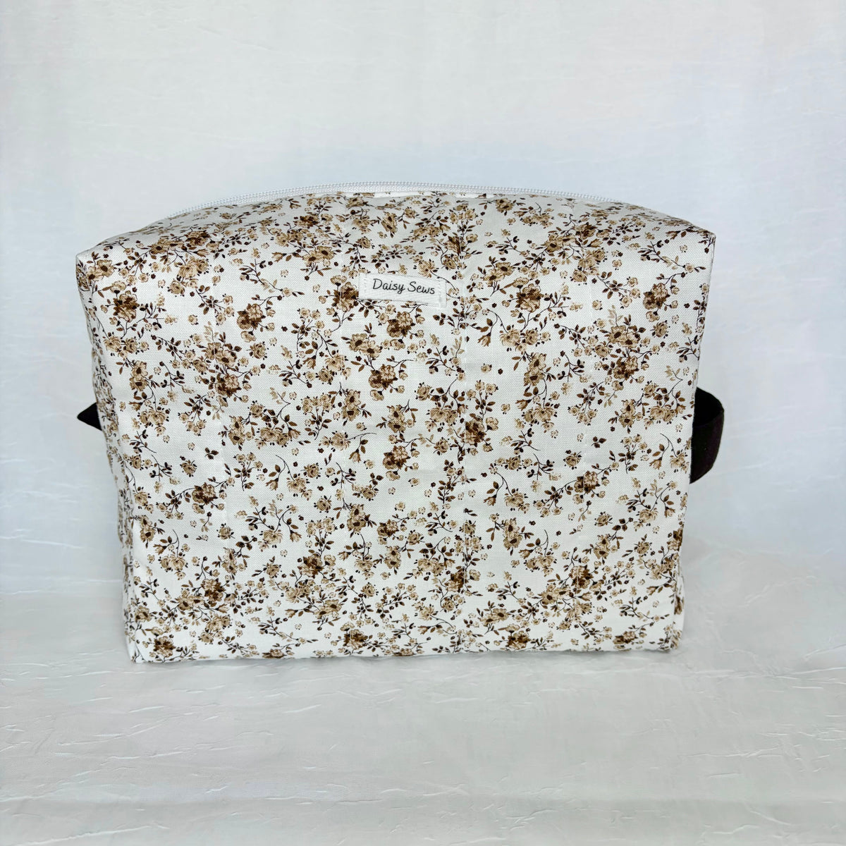 A rectangular fabric pouch or bag with a dense small floral print in brown, beige, and black on an off-white background, photographed against a light gray fabric backdrop.