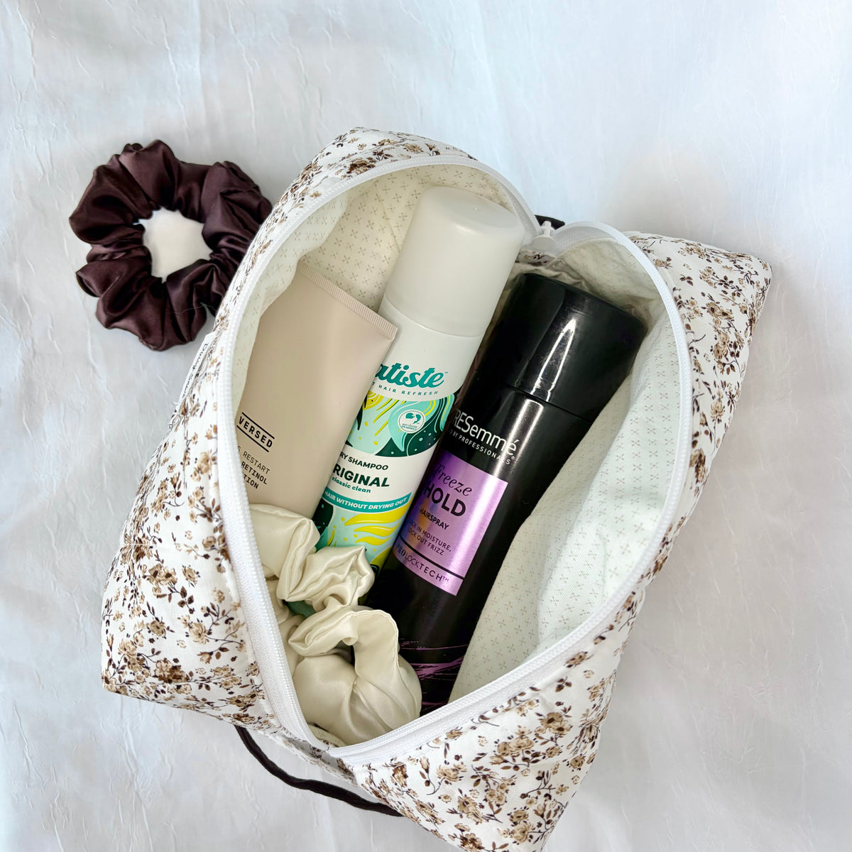 A floral-patterned zippered travel toiletry bag filled with personal care items, including a dark brown scrunchie, on a white textured fabric background.