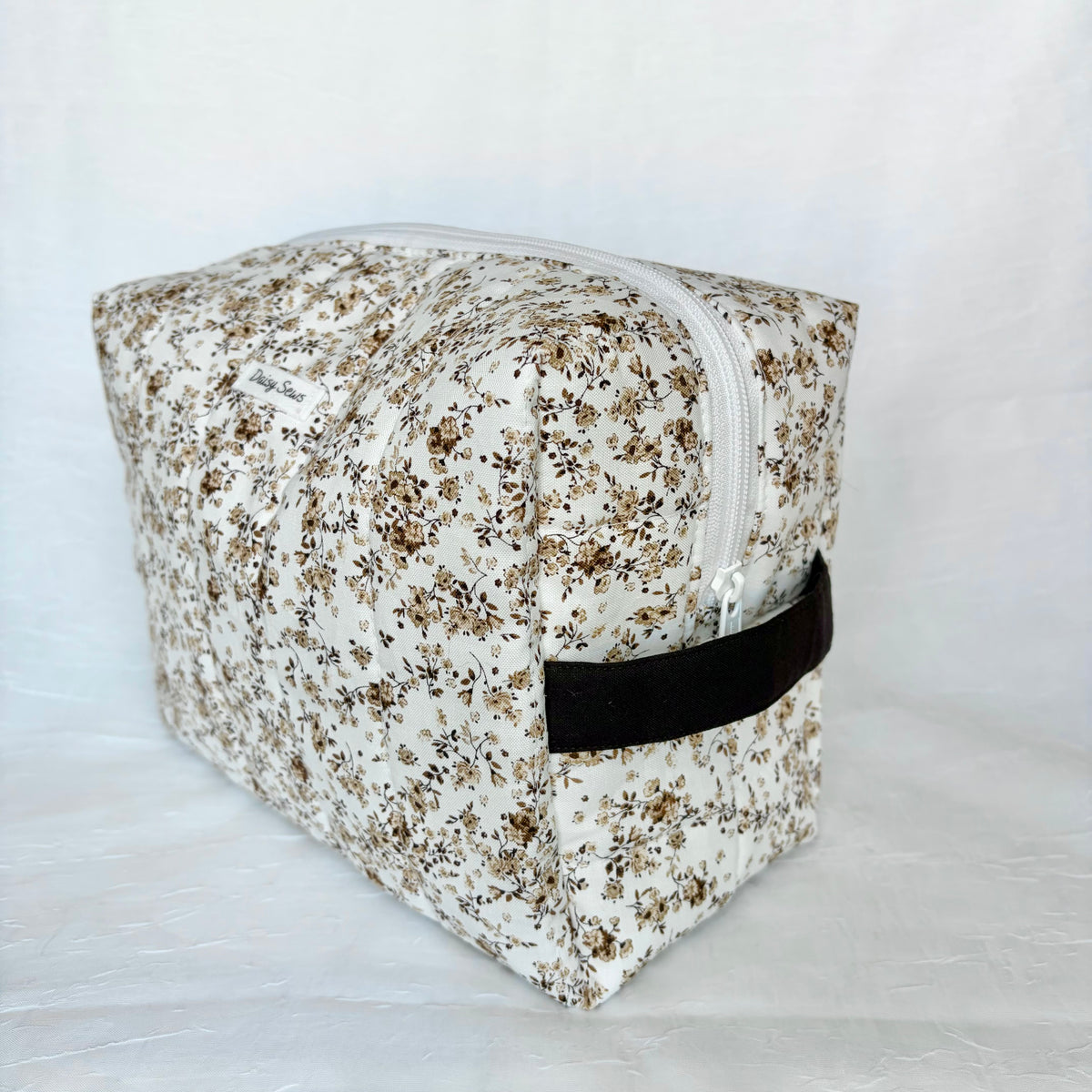 A square zippered cosmetic pouch with a white background and a dense brown and beige floral print, featuring a dark brown handle.