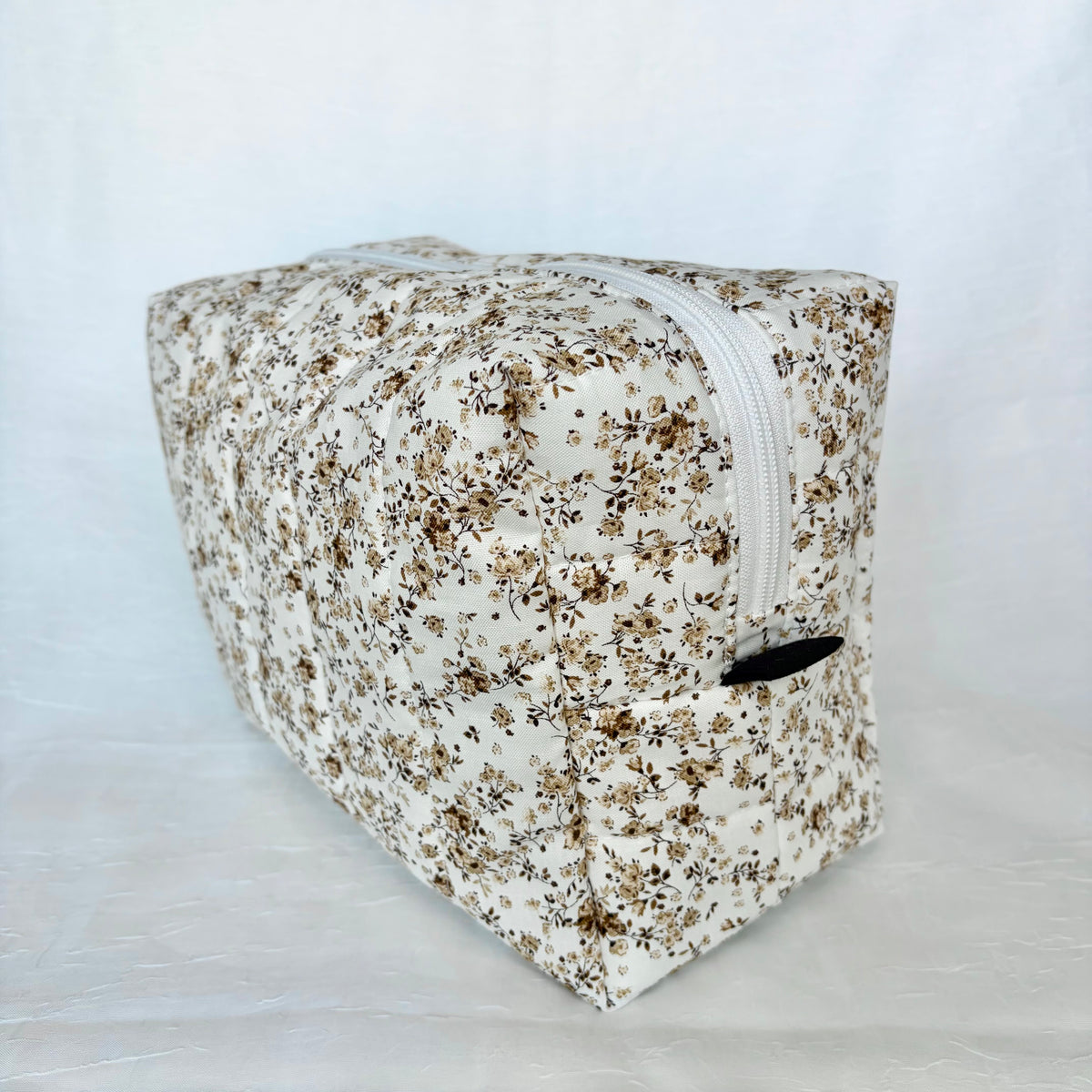 A rectangular zippered pouch with a dense brown and beige floral print on a white background, featuring a white zipper and dark brown pull tab.