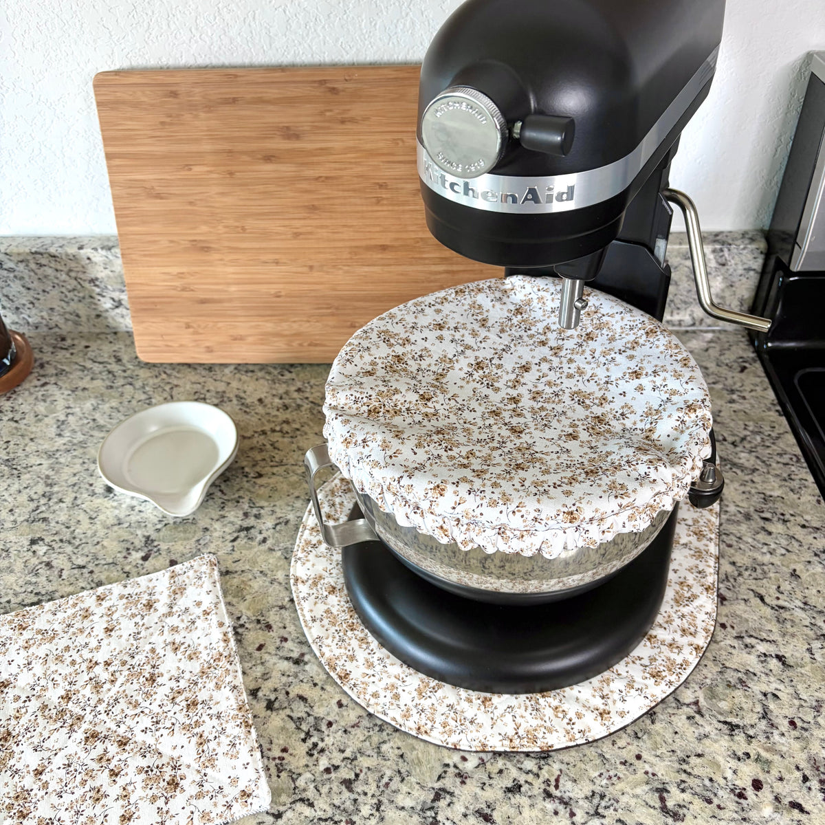 A black KitchenAid stand mixer on a granite kitchen counter, covered with a white linen tablecloth featuring a brown floral pattern. A matching patterned napkin is laid on the counter in the foreground, with a wooden cutting board and a small white dish in the background.